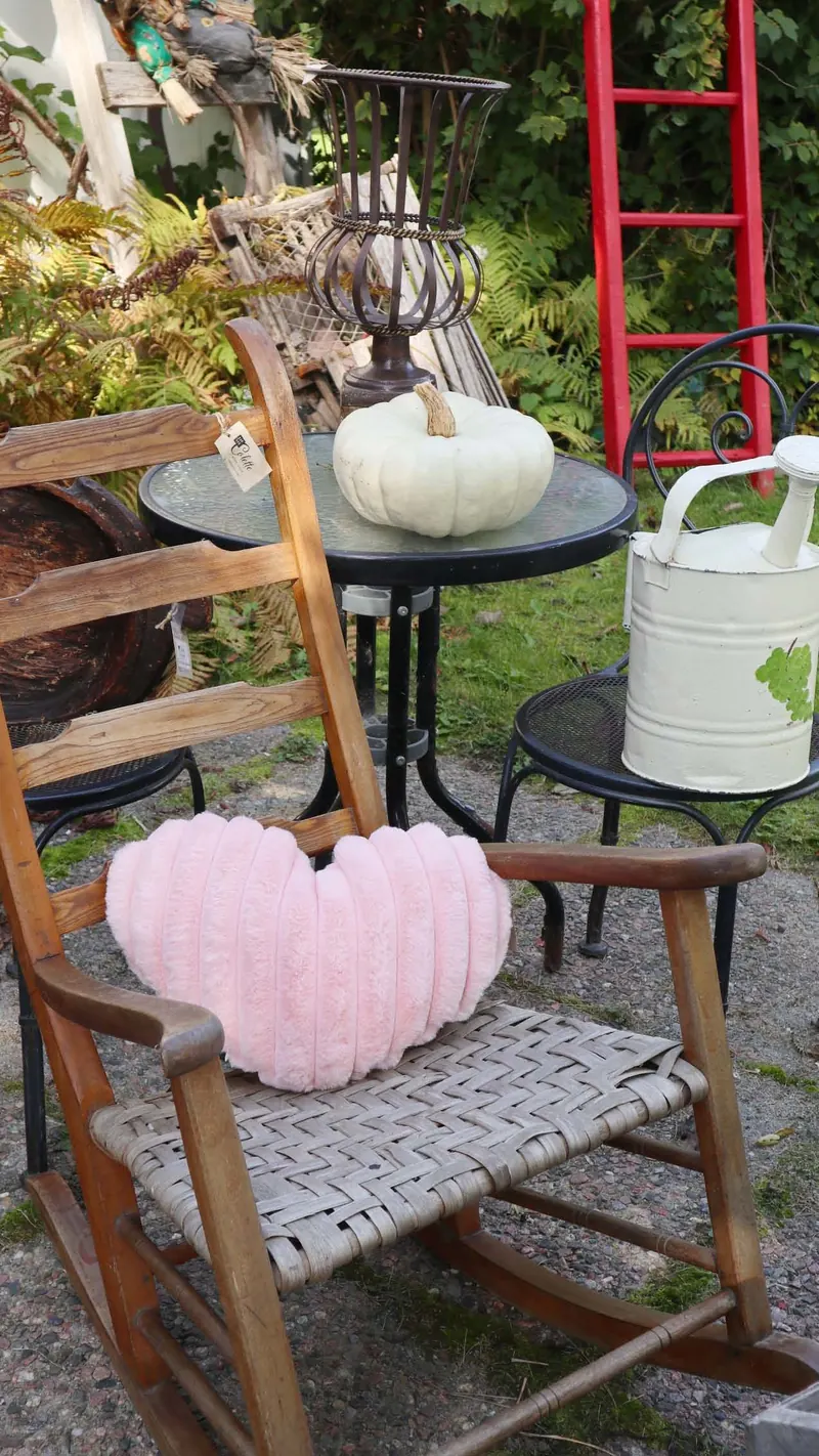 Chaise berçante ancienne en bois au siège tressé avec un coussin cœur rose, table bistro et arrosoir émaillé au jardin
